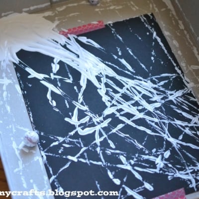 A cardboard box contains a black sheet decorated with white paint streaks made by rolling marbles through the paint, creating a marbled art pattern that resembles a spider web. The corner of a wooden table is visible in the background.