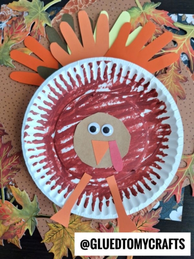 Learn how to make a paper plate turkey using a painted plate, orange and brown paper feathers, googly eyes, a red wattle, and orange legs. Surrounded by autumn leaves and dots, this paper plate turkey is perfect for fall crafts.
