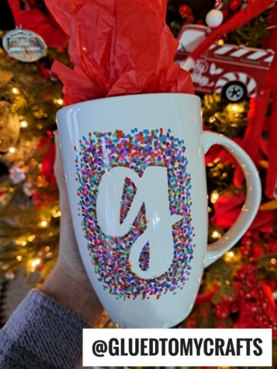 A hand holds a white DIY mug with a colorful dotted border and a large G in the center, in front of a decorated Christmas tree. Red tissue paper fills the mug, showcasing the creativity of Sharpie painted mugs. The text @GLUEDTOMYCRAFTS is at the bottom.