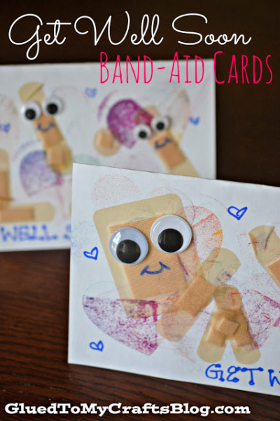 Close-up of handmade band-aid cards featuring band-aids arranged as cheerful figures with googly eyes and smiles, set against a white background with blue hearts. Text reads Get Well Soon BAND-AID CARDS.
