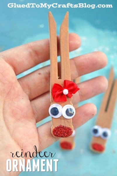 A hand holds one of the charming reindeer clothespin ornaments, featuring googly eyes, a red glittery nose, and a festive red bow. Another similar ornament appears in the blurry background.