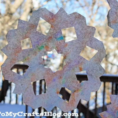 Coffee filter snowflakes with colorful patterns hang on a window, creating a cheerful winter scene. Outside, a snowy wooden deck and trees are visible.