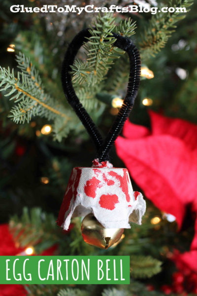 A handmade egg carton Christmas bell ornament crafted from a painted egg carton piece, decorated with red spots and a golden bell hanging below, attached by a black pipe cleaner to a tree branch.