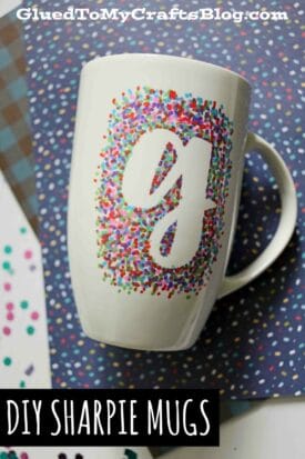A white mug decorated with colorful Sharpie dots surrounding a large white lowercase g, placed on patterned paper. Text above reads “GluedToMyCraftsBlog.com,” and below, “DIY Sharpie painted mugs.”.