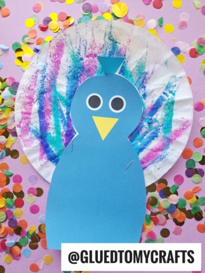 A vibrant coffee filter peacock craft made from blue paper and a decorated coffee filter in purple, blue, and pink streaks, surrounded by multicolored confetti, with the text @GLUEDTOMYCRAFTS at the bottom.