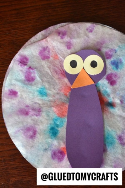A colorful owl craft made with a coffee filter for the body—just like coffee filter peacocks—decorated with purple, blue, and red dots, plus purple and orange paper for the face and body. Text reads @GLUEDTOMYCRAFTS at the bottom.