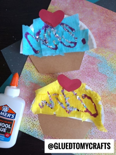 Two colorful cupcake kid crafts made from paper, tissue, and glitter glue sit on a pastel, speckled background. A bottle of Elmer’s glue is nearby. Each cupcake has a red heart on top. @GLUEDTOMYCRAFTS is labeled at the bottom.