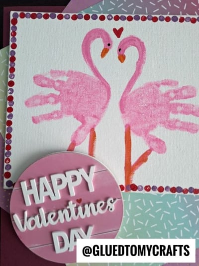 A handmade Valentine’s card features a charming handprint flamingo pair, their necks forming a heart. Beside them is a pink sign reading “Happy Valentine’s Day,” perfect inspiration for your own handprint flamingo canvas.
