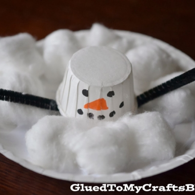 A homemade cotton ball melted snowman craft made from a small paper cup for the head, black pipe cleaners for arms, and soft cotton balls for snow, all displayed on a white paper plate.