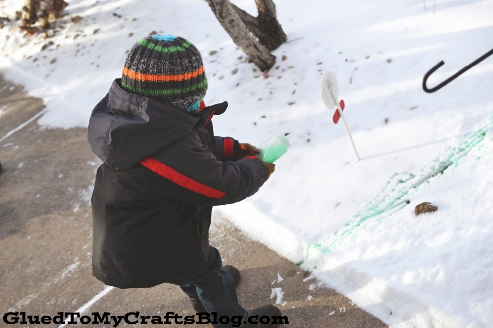 A child in a winter coat and striped hat uses snow paint, spraying green liquid onto the snow beside a sidewalk. Trees and a small sign are visible in the snowy background. The image features “GluedToMyCraftsBlog.com” at the bottom.