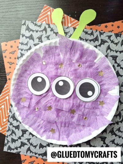 A whimsical paper plate alien craft featuring three large eyes, purple tissue paper, gold stars, and green antennae sits atop orange and black Halloween-themed paper. @GLUEDTOMYCRAFTS text is at the bottom.