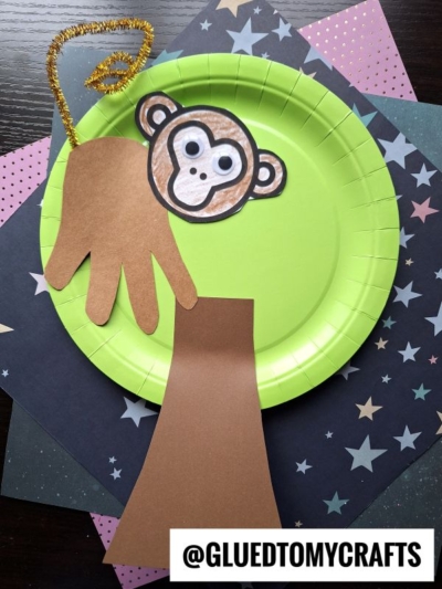 A paper plate craft featuring a handprint monkey made from brown paper, a colored monkey face, and a gold pipe cleaner tail, with star-patterned paper in the background. The @GLUEDTOMYCRAFTS watermark is visible.