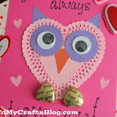 A handmade Valentines card features a heart doily owl crafted from pink and purple paper hearts, googly eyes, and gold-wrapped chocolate heart feet.
