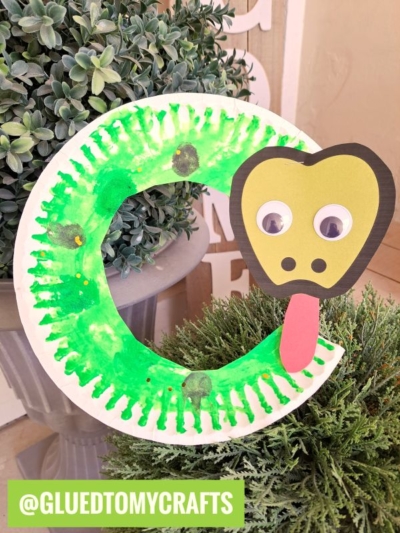 A paper plate snake craft painted green and shaped like a snake, with googly eyes and a red tongue, is displayed among green plants. The Instagram handle @gluedtomycrafts is visible at the bottom.