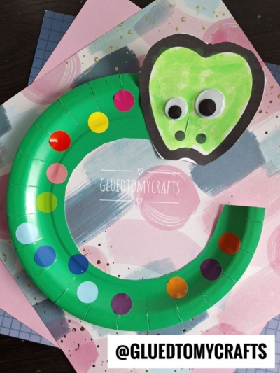 A vibrant paper plate snake with a heart-shaped head, googly eyes, and colorful dot stickers winds across pastel, patterned backgrounds— a fun and easy paper plate snake craft for kids.