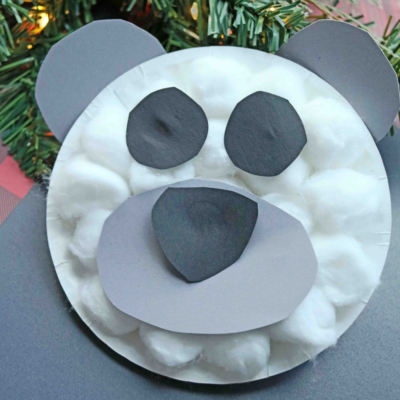 A paper plate polar bear face made with cotton balls and cut-out gray and black paper sits in front of a Christmas tree branch with red berries. Text: “Paper Plate Polar Bear.”.