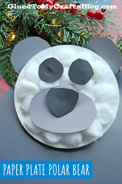 A paper plate polar bear face made with cotton balls and cut-out gray and black paper sits in front of a Christmas tree branch with red berries. Text: “Paper Plate Polar Bear.”.