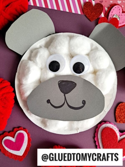 A paper plate polar bear face craft made from cotton balls and gray paper, with large googly eyes and a drawn-on nose and mouth. Red and pink felt hearts decorate the background for a cute touch.