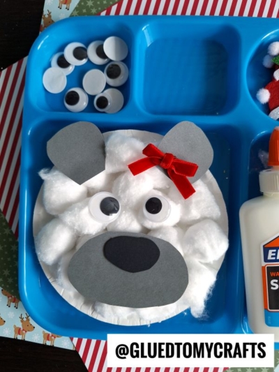 A blue tray holds a paper plate polar bear craft made from cotton balls, gray paper, googly eyes, and a red bow. Nearby are extra googly eyes, glue, and craft supplies. @GLUEDTOMYCRAFTS is written at the bottom.