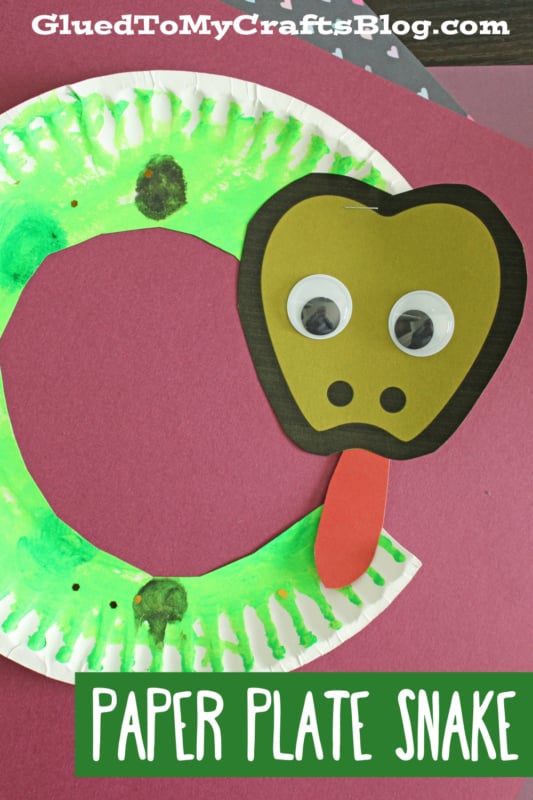 A creative paper plate snake craft featuring a brown paper head, big googly eyes, and a red tongue, all set against a purple backdrop. Text reads Paper Plate Snake and GluedToMyCraftsBlog.com.