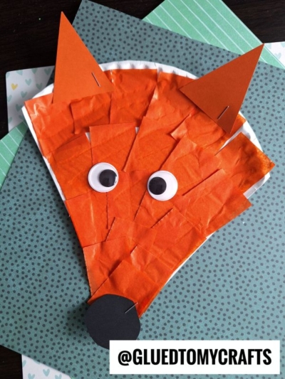 A paper plate fox face craft made with orange tissue paper, construction paper ears and nose, googly eyes, and displayed on green and white patterned layers. Perfect as a Dr. Seuss fox craft! Handle @GLUEDTOMYCRAFTS is on the bottom.