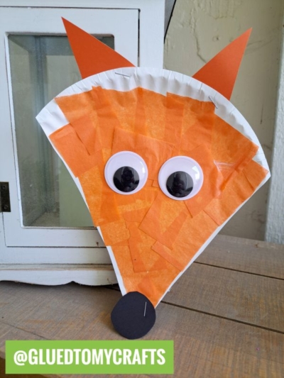 A paper plate fox craft, inspired by Dr. Seuss, features orange tissue paper, large googly eyes, triangle ears, and a black oval nose. This playful paper plate orange fox sits on a wooden surface near a white cabinet. @GLUEDTOMYCRAFTS is on a green label.