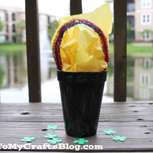 A black Styrofoam cup filled with yellow tissue paper and a rainbow made of pipe cleaners sits on a wooden surface with green paper shamrocks.
