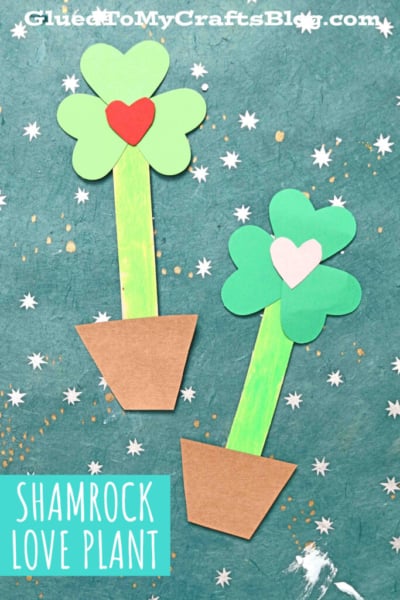 Two paper shamrock love plants in flower pots, each with a heart in the center, are displayed on a teal background with white star and dot patterns. Text at the bottom reads Shamrock Love Plant.