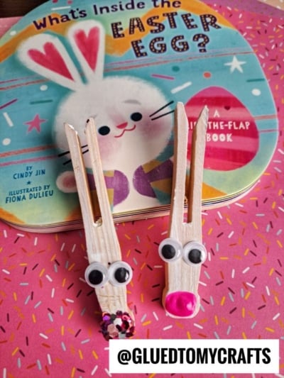 Two bunny clothespin crafts with googly eyes and pink noses sit in front of a colorful Easter book featuring a bunny illustration. A watermark reads @GLUEDTOMYCRAFTS.