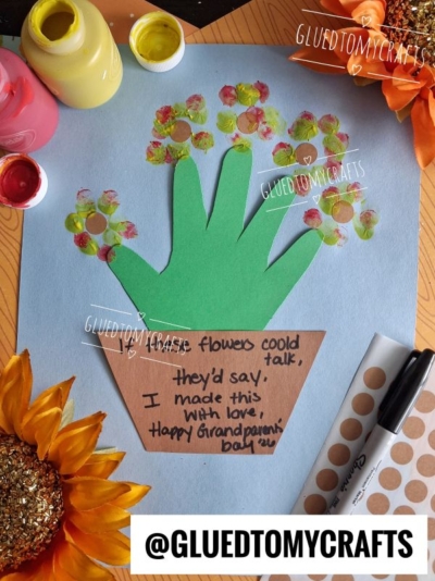 A handmade card shows a green handprint flowerpot as a plant with fingerprint flowers blooming in a pot. The pot says, If flowers could talk, they’d say, I made this with love. Happy Grandparents Day! Surrounded by art supplies, it’s the perfect keepsake to celebrate family.