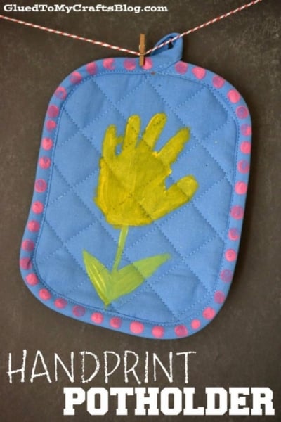 A blue quilted flower potholder with a pink polka-dot border features a yellow handprint painted to look like a flower, complete with green stem and leaves. This charming handprint flower potholder hangs by a string against a dark background.