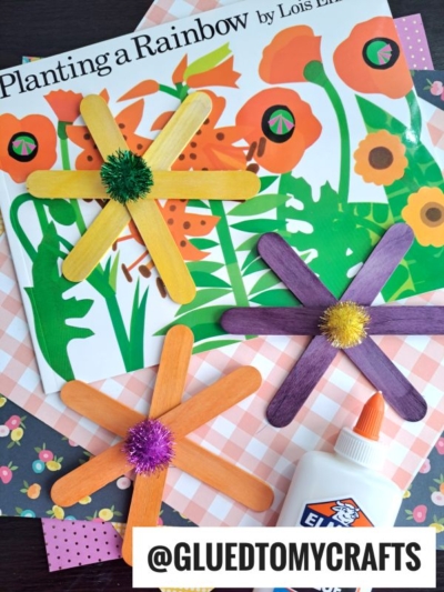 Colorful popsicle stick flowers with pom-pom centers are arranged on top of a Planting a Rainbow book, patterned paper, and a bottle of Elmer’s glue. @GLUEDTOMYCRAFTS is written at the bottom.