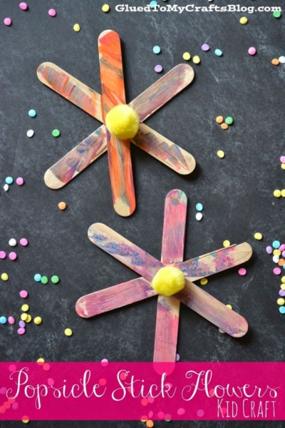 Two colorful popsicle stick flowers with yellow pom-pom centers sit on a dark background, surrounded by scattered confetti.