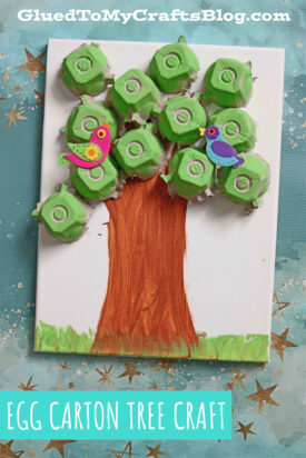 A craft project featuring an egg carton tree canvas—painted egg carton pieces form the leafy branches, with two colorful bird cutouts perched atop. The blue background is accented by shimmering gold stars.