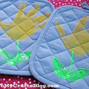 Two quilted blue handprint flower potholders with yellow designs resembling flowers, each with green leaves and stems. The potholders rest on a pink polka-dotted background. GluedToMyCraftsBlog.com is written at the bottom.