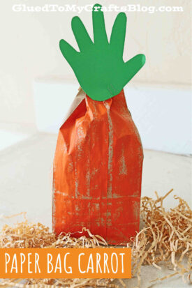 A paper bag carrot painted orange, with a green handprint cutout as its leafy top, sits on shredded paper. This fun carrot paper bag craft is labeled “PAPER BAG CARROT” in text on the image.