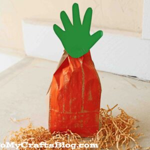 A paper bag carrot painted orange, with a green handprint cutout as its leafy top, sits on shredded paper.
