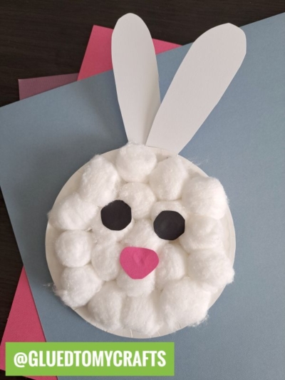 A cotton ball Easter bunny craft featuring a paper plate covered in fluffy cotton balls, two paper ears, black eyes, and a pink nose. Blue and pink paper sheets form the background. @GLUEDTOMYCRAFTS is labeled at the bottom.