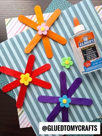 Colorful craft stick flowers with felt centers are arranged on striped and polka dot paper backgrounds, next to a bottle of Elmer’s clear glue. The image includes the text @GLUEDTOMYCRAFTS at the bottom. Perfect for popsicle stick flowers crafts!.
