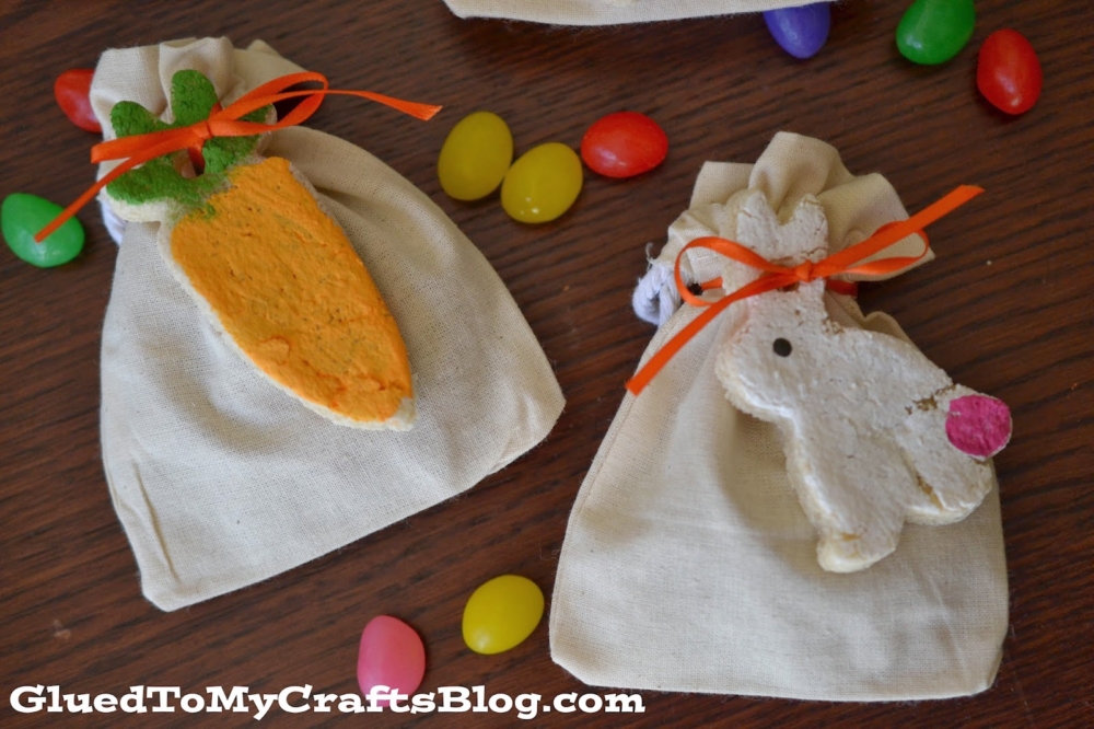 Two small drawstring cloth bags with orange ribbons are decorated, one with a carrot-shaped salt dough Easter tag and the other with a bunny face ornament. Colorful jellybeans are scattered on the wooden surface.