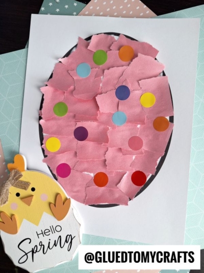 A torn paper Easter egg craft featuring pink paper pieces and colorful dot stickers on white paper, displayed beside a wooden chick cutout that says "Hello Spring" with @GLUEDTOMYCRAFTS at the bottom.