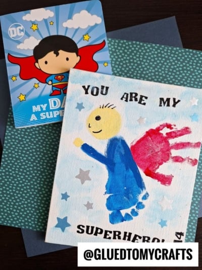 A handmade superhero card with a blue footprint and red handprint cape, next to a cartoon card of a superhero dad. Text reads “You are my superhero.” Perfect for gifting or displaying as your own 'my superhero canvas.' Background is decorated in blue, green, and white.
