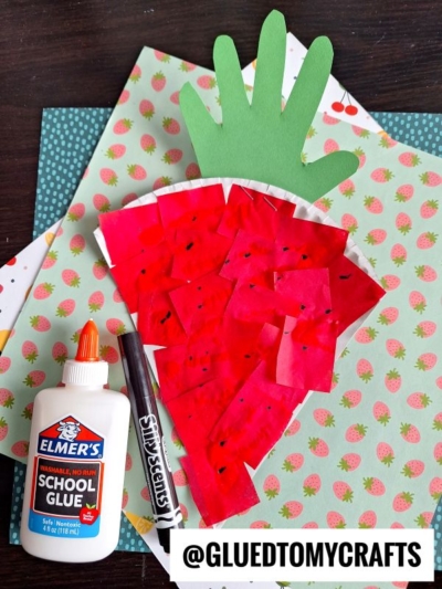 A handmade carrot craft made with red tissue paper squares glued onto a semicircle, similar to making a paper strawberry, topped with a green paper hand cutout as leaves. Elmers glue, a black marker, and colorful paper are nearby.