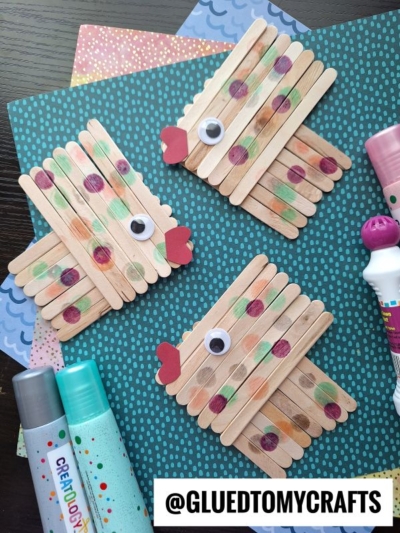 Three craft stick fish, also known as Popsicle Stick Fish, are decorated with painted spots, red paper mouths, and googly eyes. They’re displayed on a teal patterned background with markers and paint daubers nearby.