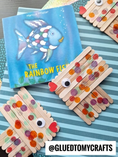 A childrens book titled The Rainbow Fish is displayed next to craft stick fish crafts made from popsicle sticks, painted with colorful dots, googly eyes, and red heart-shaped mouths on a striped background. @gluedtomycrafts watermark present.