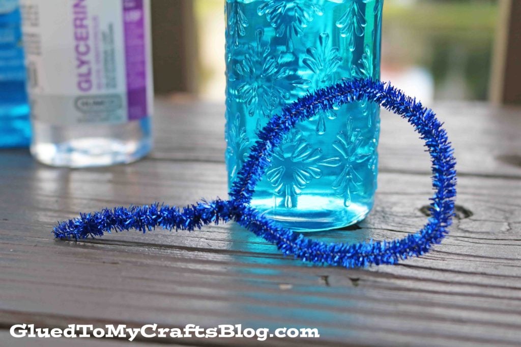 A blue pipe cleaner bent into a bubble wand shape lies on a wooden surface near a blue patterned bottle of homemade bubble mixture and a clear glycerin bottle. Text at the bottom reads GluedToMyCraftsBlog.com.