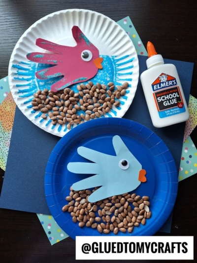 Two paper plates each display a handprint fishbowl craft—fish made from cut-out hand shapes with googly eyes and paper mouths swim above a layer of dried beans, next to a bottle of glue. @GLUEDTOMYCRAFTS is visible at the bottom.