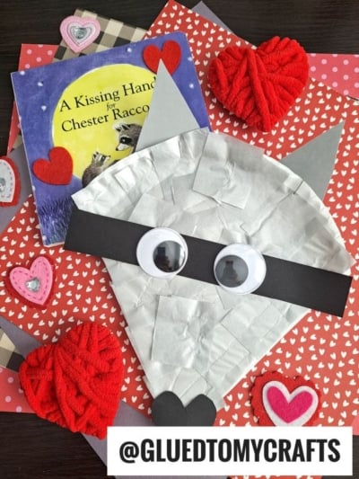 A raccoon face made from gray paper, googly eyes, and heart shapes sits on patterned paper with red and pink hearts, alongside the book A Kissing Hand for Chester Raccoon. Perfect inspiration for a cute raccoon paper plate craft! Username @GLUEDTOMYCRAFTS is visible.