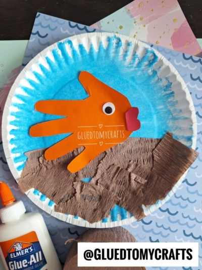 A handmade handprint fishbowl craft on a paper plate, featuring a blue painted background, brown paper sand, and an orange handprint fish. Elmer’s glue and craft papers are beside this creative Paper Plate Fish bowl project.