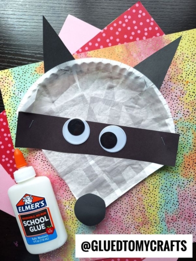 A paper plate raccoon craft, complete with black paper ears, a black mask, and googly eyes, sits on colorful paper. A bottle of Elmer’s school glue is next to it, alongside the text @GLUEDTOMYCRAFTS.